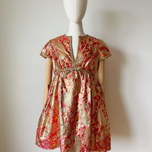 Może przedstawiać: Mini sukienka w stylu vintage z czerwonej i złotej tkaniny brokatowej. Sukienka ma dekolt w kształcie litery V, krótkie rękawy i dopasowaną talię. Tkanina ma wzór paisley. Sukienka jest prezentowana na manekinie.