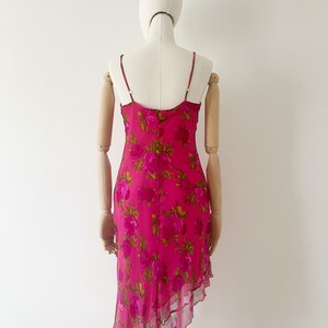 K&ouml;nnte beinhalten: Ein leuchtend pinkfarbenes Slip-Kleid mit einem floralen Rosenmuster in Pink- und Gr&uuml;nt&ouml;nen. Das Kleid hat d&uuml;nne Spaghettitr&auml;ger und einen asymmetrischen Saum mit R&uuml;schenbesatz. Das Kleid wird auf einer Schaufensterpuppe pr&auml;sentiert.