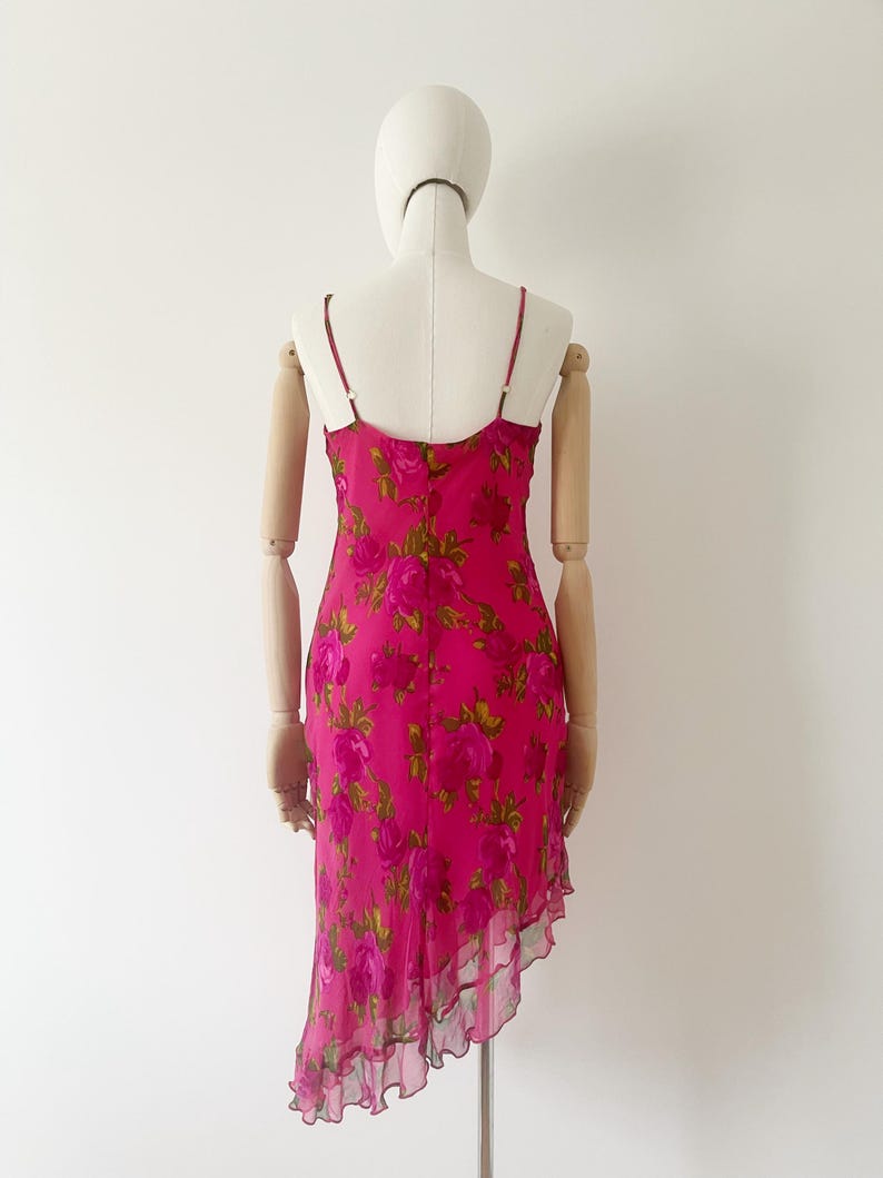 K&ouml;nnte beinhalten: Ein leuchtend pinkfarbenes Slip-Kleid mit einem floralen Rosenmuster in Pink- und Gr&uuml;nt&ouml;nen. Das Kleid hat d&uuml;nne Spaghettitr&auml;ger und einen asymmetrischen Saum mit R&uuml;schenbesatz. Das Kleid wird auf einer Schaufensterpuppe pr&auml;sentiert.