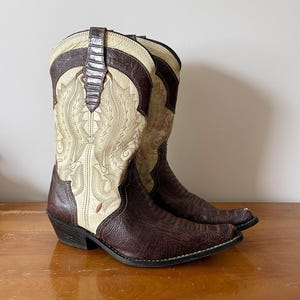 May include: A pair of cowboy boots with a cream-colored shaft featuring intricate embroidery. The boots have a dark brown, textured leather foot and accents, with a brown leather strap at the top. The boots are set on a wooden surface.