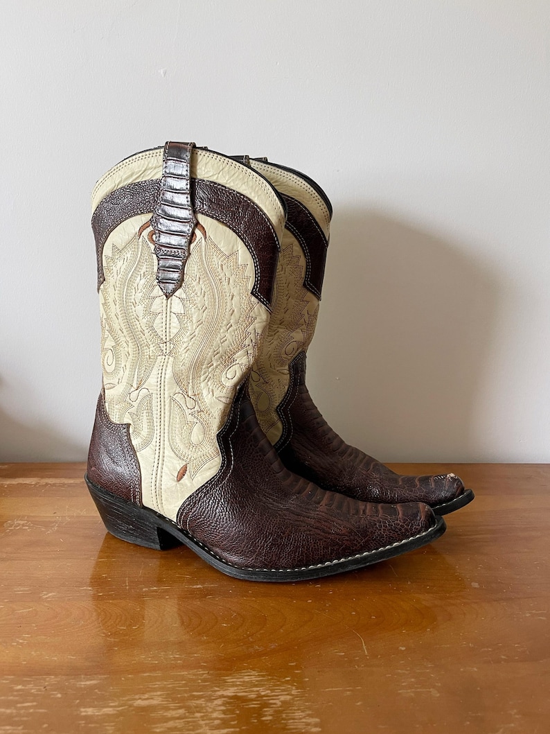 May include: A pair of cowboy boots with a cream-colored shaft featuring intricate embroidery. The boots have a dark brown, textured leather foot and accents, with a brown leather strap at the top. The boots are set on a wooden surface.