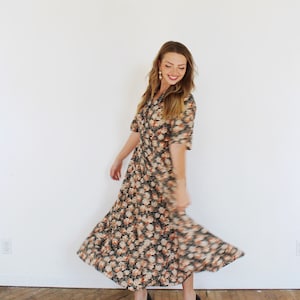 K&ouml;nnte beinhalten: Eine Frau tr&auml;gt ein langes, blumiges Kleid mit dunkelgr&uuml;nem Hintergrund und orangefarbenen und wei&szlig;en Blumen. Sie tr&auml;gt schwarze Schuhe mit Absatz und hat lange blonde Haare.