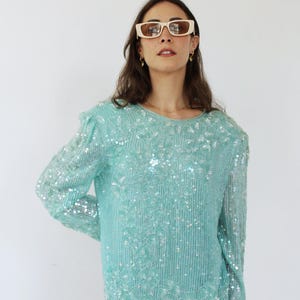Puede incluir: Una blusa turquesa con lentejuelas, mangas largas y cuello redondo. La blusa tiene un patrón floral y está hecha de una tela transparente.