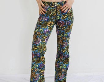 May include: Floral print pants with a blue background and a variety of colorful flowers. The pants have a button closure and a flared leg. The model is wearing a gray crop top and tan shoes.