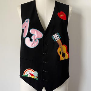 May include: A black button-up vest featuring several colorful embroidered patches. The patches include a large pink number thirteen, a yellow acoustic guitar, a red heart with the text "SMAK!", and a landscape scene with a rainbow and house.