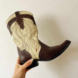 May include: A brown and cream-colored cowboy boot with intricate embroidered detailing. The boot features a pointed toe and a stacked heel. The upper part of the boot has a cream-colored design with brown accents.