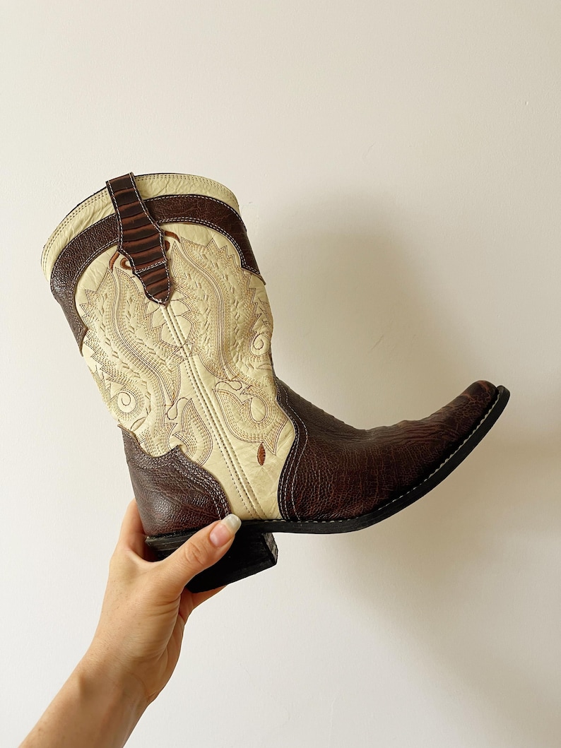 May include: A brown and cream-colored cowboy boot with intricate embroidered detailing. The boot features a pointed toe and a stacked heel. The upper part of the boot has a cream-colored design with brown accents.