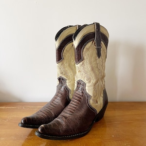 May include: A pair of cowboy boots with a brown, textured vamp and a cream-colored shaft with decorative stitching. The boots have a pointed toe and a stacked heel, perfect for a Western-themed outfit.