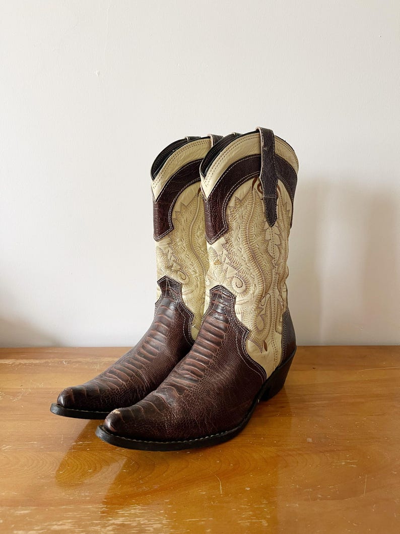 May include: A pair of cowboy boots with a brown, textured vamp and a cream-colored shaft with decorative stitching. The boots have a pointed toe and a stacked heel, perfect for a Western-themed outfit.