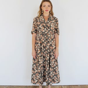 K&ouml;nnte beinhalten: Ein langes, blumiges Kleid mit Kragen und kurzen &Auml;rmeln. Das Kleid hat einen taillierten Oberteil und einen flie&szlig;enden Rock. Das Model tr&auml;gt schwarze Abs&auml;tze.