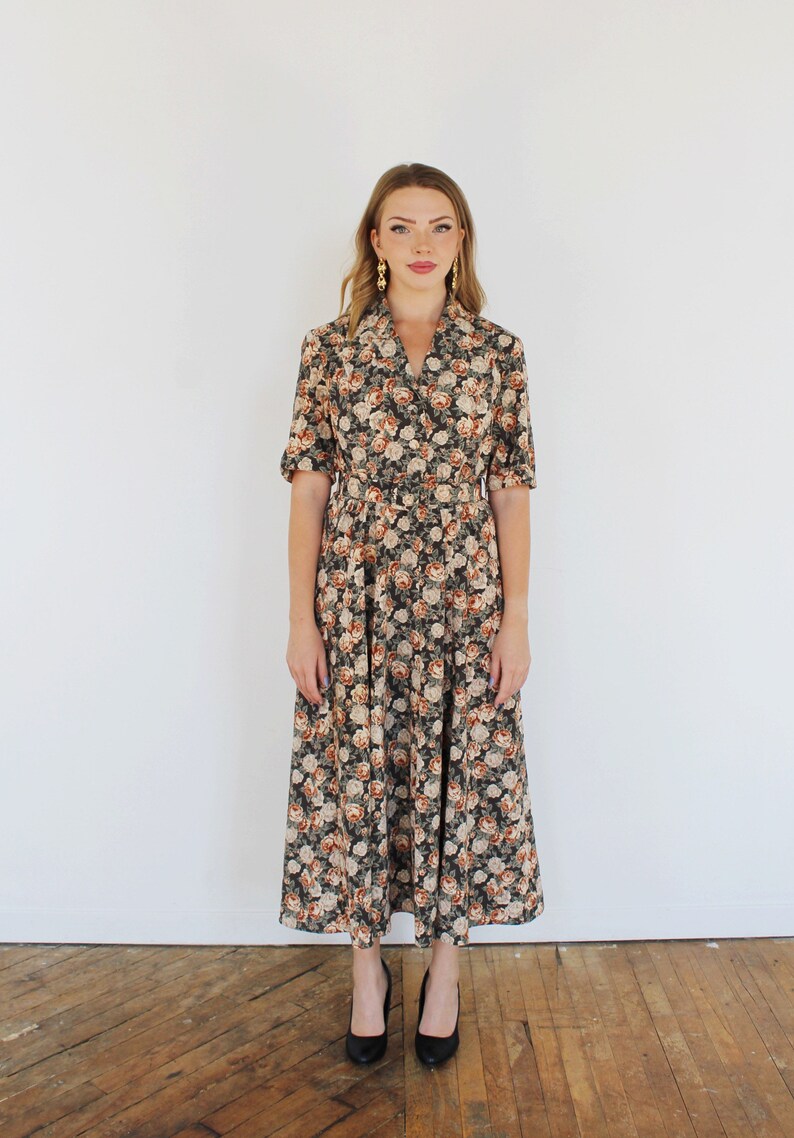 K&ouml;nnte beinhalten: Ein langes, blumiges Kleid mit Kragen und kurzen &Auml;rmeln. Das Kleid hat einen taillierten Oberteil und einen flie&szlig;enden Rock. Das Model tr&auml;gt schwarze Abs&auml;tze.