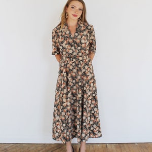K&ouml;nnte beinhalten: Ein langes, schwarzes Blumenkleid mit V-Ausschnitt und G&uuml;rtel. Das Kleid hat ein Blumenmuster mit rosa, wei&szlig;en und orangefarbenen Blumen. Das Model tr&auml;gt schwarze Abs&auml;tze.