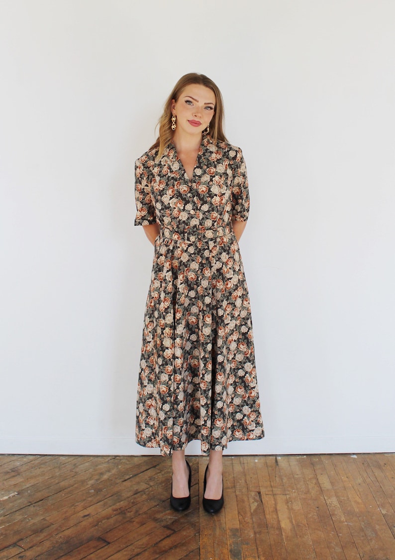 K&ouml;nnte beinhalten: Ein langes, schwarzes Blumenkleid mit V-Ausschnitt und G&uuml;rtel. Das Kleid hat ein Blumenmuster mit rosa, wei&szlig;en und orangefarbenen Blumen. Das Model tr&auml;gt schwarze Abs&auml;tze.