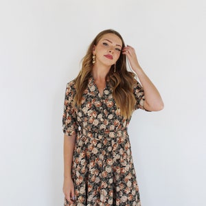 K&ouml;nnte beinhalten: Eine Frau tr&auml;gt ein dunkelgr&uuml;nes Blumenkleid mit einem braunen G&uuml;rtel. Das Kleid hat einen V-Ausschnitt und kurze &Auml;rmel. Sie tr&auml;gt goldene Ohrringe.