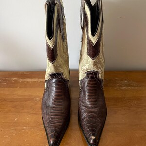 May include: A pair of cowboy boots with a pointed toe. The boots have a brown, textured vamp and a cream-colored shaft with decorative stitching. The top of the shaft is brown. The boots are made of leather.