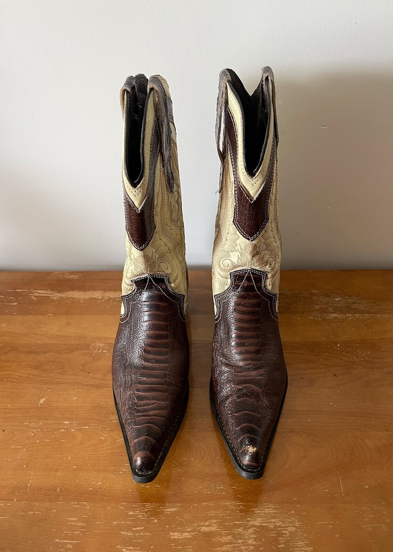 May include: A pair of cowboy boots with a pointed toe. The boots have a brown, textured vamp and a cream-colored shaft with decorative stitching. The top of the shaft is brown. The boots are made of leather.