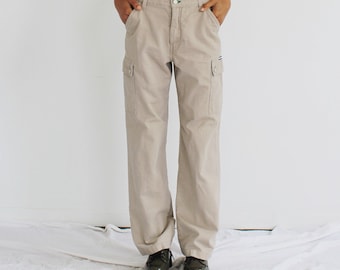 Könnte beinhalten: Beigefarbene Cargohose mit mehreren Taschen und lockerer Passform. Die Hose hat einen Knopf- und Reißverschluss und wird mit dunkelgrünen Schuhen getragen. Das Model trägt ein hautfarbenes Oberteil.