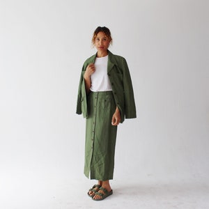 Puede incluir: Una mujer con una falda verde de botones y una chaqueta a juego. Lleva una camiseta blanca debajo de la chaqueta y sandalias marrones.