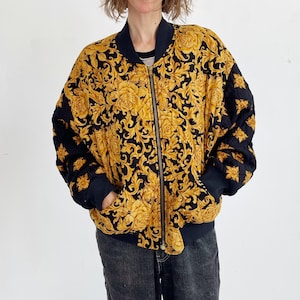 Puede incluir: Chaqueta bomber negra y dorada con un diseño floral. La chaqueta tiene cierre de cremallera y puños y cuello negros. El diseño floral dorado está sobre un fondo negro. La chaqueta se lleva sobre una camiseta negra y vaqueros azules.