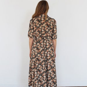 K&ouml;nnte beinhalten: Ein langes, schwarz-braunes Blumenkleid mit G&uuml;rtel. Das Kleid hat einen hohen Ausschnitt und kurze &Auml;rmel.