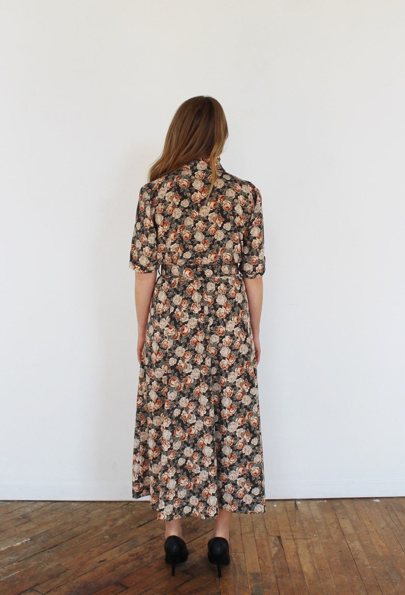 K&ouml;nnte beinhalten: Ein langes, schwarz-braunes Blumenkleid mit G&uuml;rtel. Das Kleid hat einen hohen Ausschnitt und kurze &Auml;rmel.
