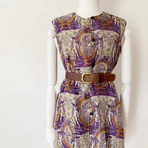 Puede incluir: Un vestido sin mangas con botones con estampado morado y dorado que presenta un patrón repetido de figuras en marcos. El vestido tiene un cinturón de cuero marrón.