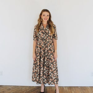 K&ouml;nnte beinhalten: Eine Frau tr&auml;gt ein langes, blumiges Kleid mit Kragen und kurzen &Auml;rmeln. Das Kleid hat einen taillierten Oberteil und einen flie&szlig;enden Rock. Sie tr&auml;gt schwarze Schuhe mit Absatz.