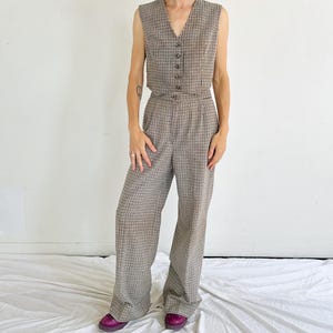 May include: A two-piece outfit comprising a sleeveless waistcoat and wide-leg trousers. The set is patterned with a brown and white check design. The waistcoat has a V-neck and button closure. The trousers have a high waist and a relaxed fit. The model is wearing purple shoes.