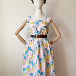 Puede incluir: Vestido patchwork vintage con cuadrados rosas, azules y amarillos, cinturón marrón y gorra con estampado floral. El vestido tiene cuello cuadrado y bolsillos. El maniquí está sobre un fondo blanco.