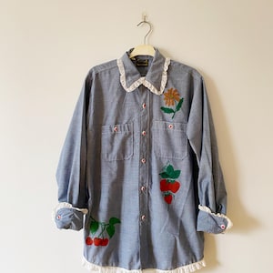 Peut inclure: Une chemise en jean bleu clair avec une bordure en dentelle blanche autour du col, des poignets et de l'ourlet. La chemise présente des motifs de fruits et de fleurs brodés en rouge, vert et jaune.