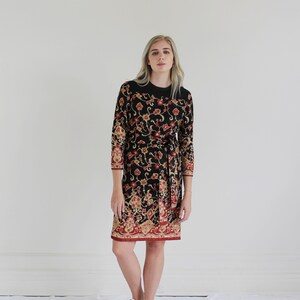 Op de afbeelding: Een zwarte jurk met een bloemenprint in tinten rood, goud en bruin. De jurk heeft lange mouwen en een strik in de taille. Het model draagt witte sneakers.