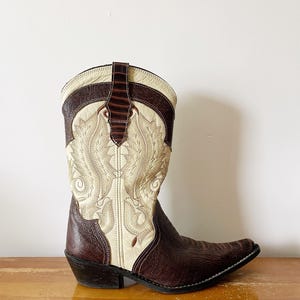 May include: A brown and cream-colored cowboy boot with intricate stitching and a pointed toe. The boot features a decorative design on the front and a dark brown leather accent at the top. The heel is approximately 2 inches high.