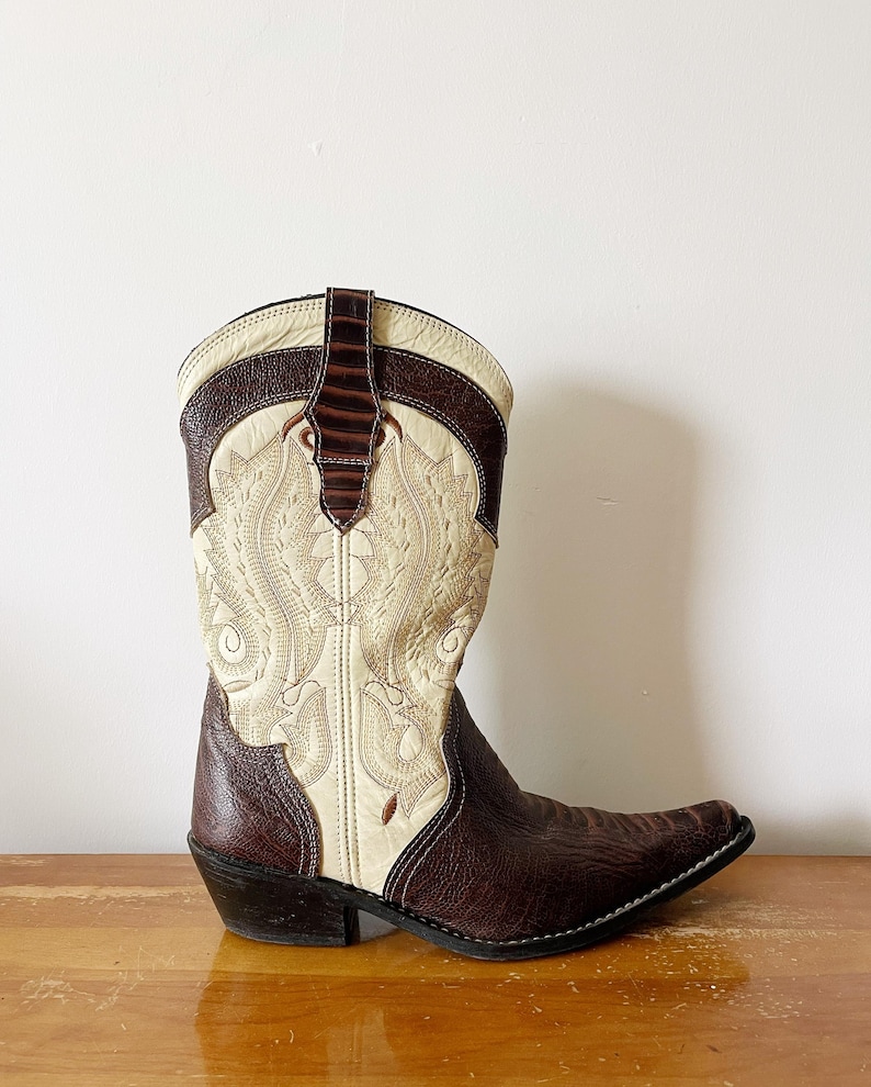 May include: A brown and cream-colored cowboy boot with intricate stitching and a pointed toe. The boot features a decorative design on the front and a dark brown leather accent at the top. The heel is approximately 2 inches high.
