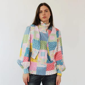 May include: A woman wearing a patchwork vest and a long-sleeved shirt with a floral pattern. The vest and shirt are both made of a light fabric and have a variety of colors, including pink, blue, green, and yellow. The woman is also wearing blue jeans.