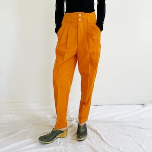 May include: A person wearing a black long-sleeved top and high-waisted, orange trousers with pleats. The trousers are paired with dark green clogs with wooden heels. The outfit is set against a white background.