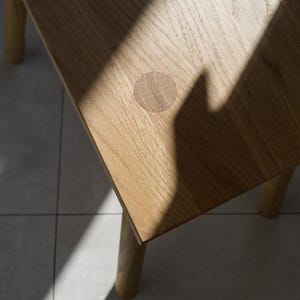 May include: Close-up of a light-colored wooden table with a round, inlaid detail. The table has four legs and a square top. A shadow from a window or other light source is cast across the table's surface.