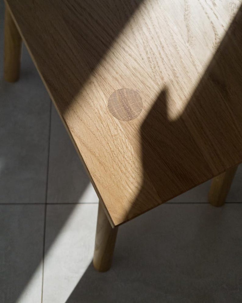 May include: Close-up of a light-colored wooden table with a round, inlaid detail. The table has four legs and a square top. A shadow from a window or other light source is cast across the table's surface.