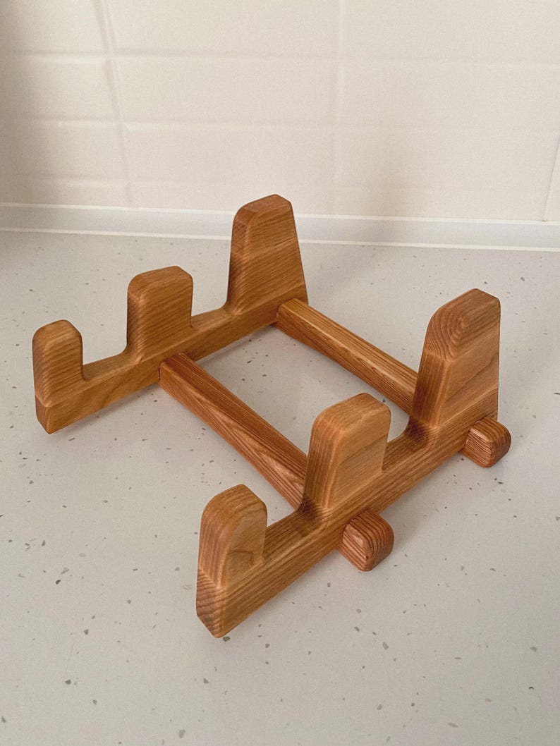 May include: A light brown wooden stand with a rectangular base and four upright supports. The stand is made of natural wood and has a simple, functional design. The stand is likely used to hold a laptop or other electronic device.