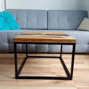 May include: A square coffee table with a wooden top and a black metal frame. The top has a dark gray epoxy river running through the center.
