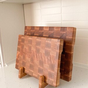 May include: Two wooden cutting boards with a butcher block design. The boards are displayed on a wooden stand. The wood grain is visible, with a mix of light and dark brown tones. The boards are rectangular.