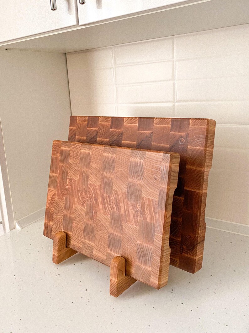 May include: Two wooden cutting boards with a butcher block design. The boards are displayed on a wooden stand. The wood grain is visible, with a mix of light and dark brown tones. The boards are rectangular.