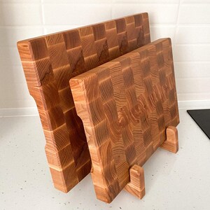 May include: Two end-grain wooden cutting boards. The boards are made of light brown wood with a checkerboard pattern. The boards are rectangular with a handle on one side and are propped up on small wooden stands.