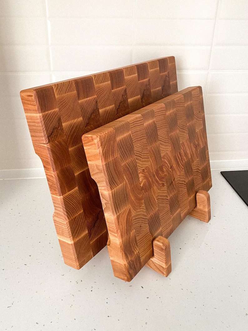 May include: Two end-grain wooden cutting boards. The boards are made of light brown wood with a checkerboard pattern. The boards are rectangular with a handle on one side and are propped up on small wooden stands.