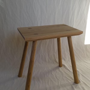 May include: A small, natural wood stool with a square seat and four angled legs. The stool is made of light-colored wood and has a simple, minimalist design. The seat has a slightly rough edge and is approximately 12 inches wide.