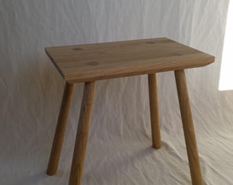 May include: A small, natural wood stool with a square seat and four angled legs. The stool is made of light-colored wood and has a simple, minimalist design. The seat has a slightly rough edge and is approximately 12 inches wide.