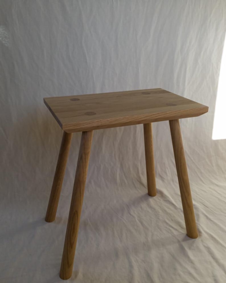 May include: A small, natural wood stool with a square seat and four angled legs. The stool is made of light-colored wood and has a simple, minimalist design. The seat has a slightly rough edge and is approximately 12 inches wide.