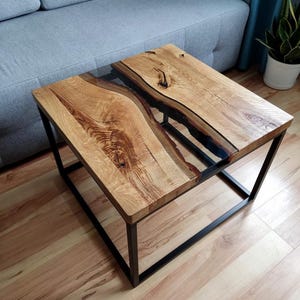 May include: A square coffee table with a black metal frame and a wooden top. The top features a clear epoxy river running through the center of the wood.
