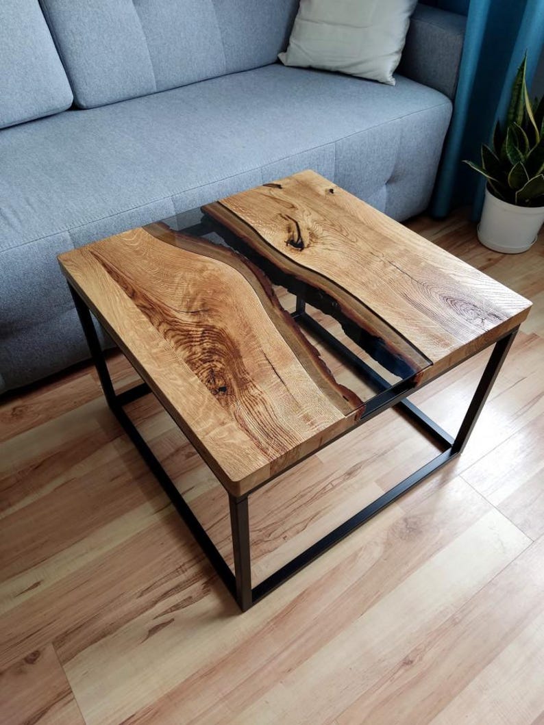 May include: A square coffee table with a black metal frame and a wooden top. The top features a clear epoxy river running through the center of the wood.