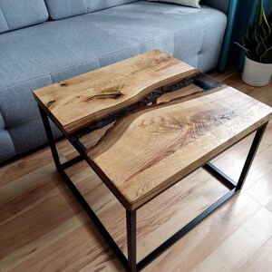 May include: A square coffee table with a wooden top and a black metal frame. The top has a natural wood grain and a clear epoxy resin inlay that resembles a river.
