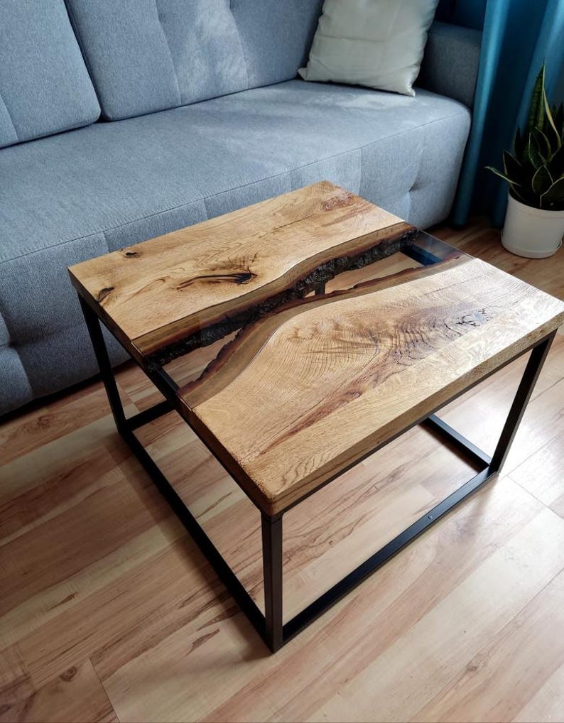 May include: A square coffee table with a wooden top and a black metal frame. The top has a natural wood grain and a clear epoxy resin inlay that resembles a river.